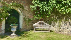 Photo of a wooden bench in the corner of a walled garden on green grass with green leaves hanging overhead. 